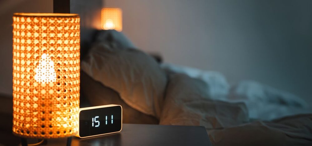 A dim bedside lamp and a glowing alarm clock at night